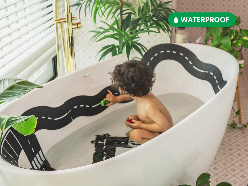 A young child enjoys creative play in the tub with toy cars and the Large Flexible Toy Road Set - Highway (24-Piece) sticker. Surrounded by green plants, a "WATERPROOF" label appears in the top right corner.