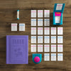 The Board Game: Taboo Vintage Bookshelf Edition is displayed on a wooden table, including a purple book-shaped box, timer, buzzer, card holder, and neatly arranged game cards.
