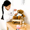 A young child dressed in a white coat and ID badge examines a teddy bear on a wooden table, using the Wooden Dentist Set for playful and imaginative role play. Toy dental tools add to the bright, creative scene.