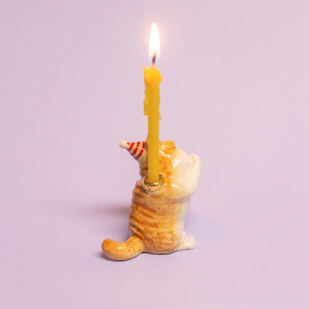 The Exotic Shorthair Cat Cake Topper features a small ceramic cat in a party hat holding a lit yellow candle—ideal as a cake decoration for cat-themed parties—shown on a plain light purple background.