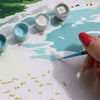 A hand with red nails paints the "Paint By Number Kit - Picnic in Étretat by Ananya Bhandari," featuring blue and white Normandy coast scenes. Open paint pots surround the canvas, showing numbered sections ready to be filled.