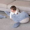 A young child in white clothes lies on the Ride-On Whale Pouf, a large plush toy with a playful whale design, placed on a carpet next to a light-colored sofa.
