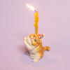 The Exotic Shorthair Cat Cake Topper is a ceramic orange and white figurine with a red party hat, holding a lit yellow candle—ideal for cat-themed parties and cake decoration, shown on a light purple background.