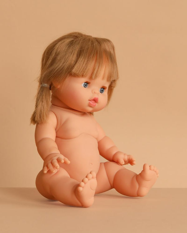 The Minikane Baby Doll (13") - Yzé sits upright on a beige surface with brown pigtails, blue eyes, and pink lips. Made in Spain, this anatomically correct doll comes unclothed; clothing sold separately.