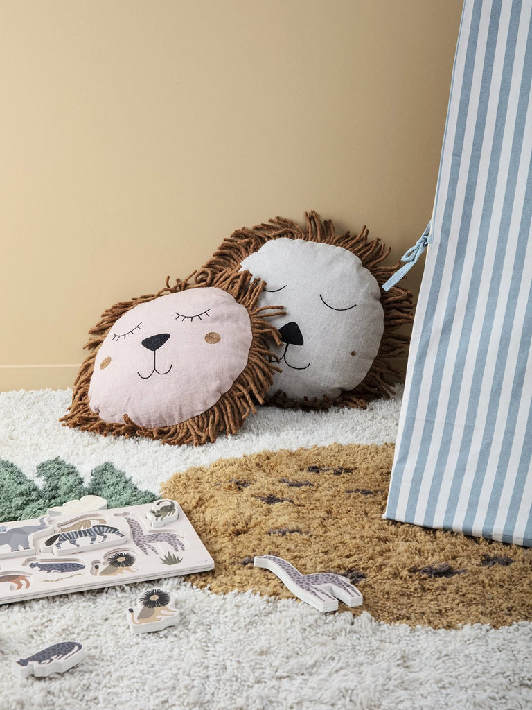 Two Ferm Living Safari Cushion - Lion pillows with sleepy faces rest against a beige wall; nearby, a blue and white striped tent and animal puzzle pieces are scattered on a fluffy rug with green leaf and brown circle patterns.