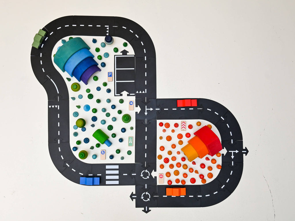 The Large Flexible Toy Road Set - Highway (24-Piece) features curved black roads with white markings, bordered by colorful wooden blocks and trees. Made from child-safe materials, it’s split into blue-green and red-orange-yellow play zones on a white base.