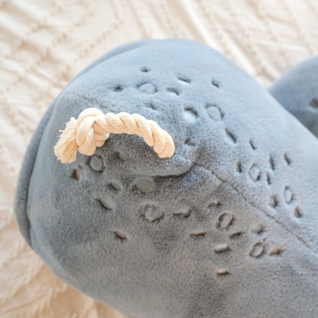A detailed close-up of the Ride-On Whale Pouf shows its soft blue plush body with textured spots, knotted rope tail, and stitched accents, resting on patterned white fabric and capturing a playful whale-inspired charm.