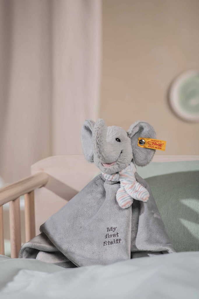 The “My First Steiff” Ellie Elephant Security Blanket Baby Toy, 10 Inches, featuring a plush gray elephant with a striped scarf and yellow tag, sits on a bed in a cozy nursery with neutral bedding and a crib in the background.