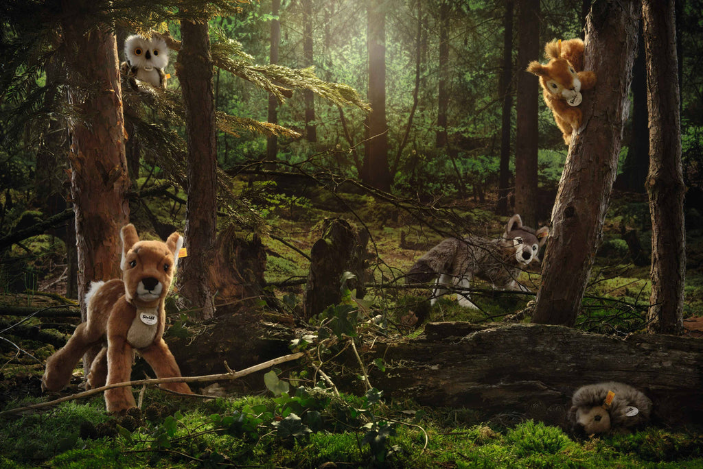 Five handmade plush toy animals, including the Steiff Joggi Hedgehog Plush Toy (6 Inches), are posed among trees and moss in a sunlit forest: two climbing, one standing, one lying behind a log, and one resting on the moss.