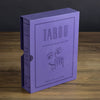 A purple box of "Board Game: Taboo Vintage Bookshelf Edition" stands upright on a wooden surface, featuring a minimalist illustration of a face with a hand partially covering its mouth.
