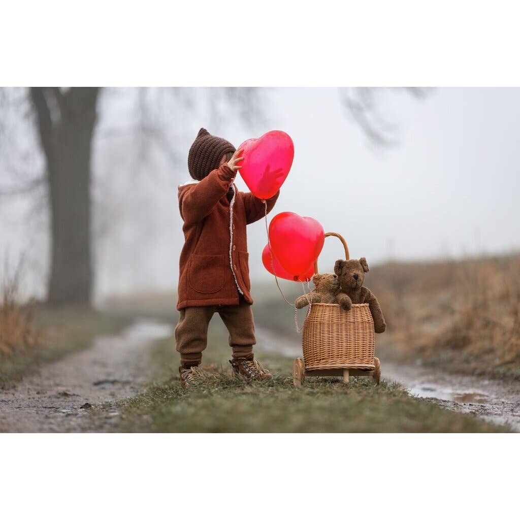 A young child in a brown coat and hat holds two red heart-shaped balloons on a misty path, beside a wicker basket with the GOTS-certified Senger Naturwelt Stuffed Animal - Large Brown Bear and an attached balloon.