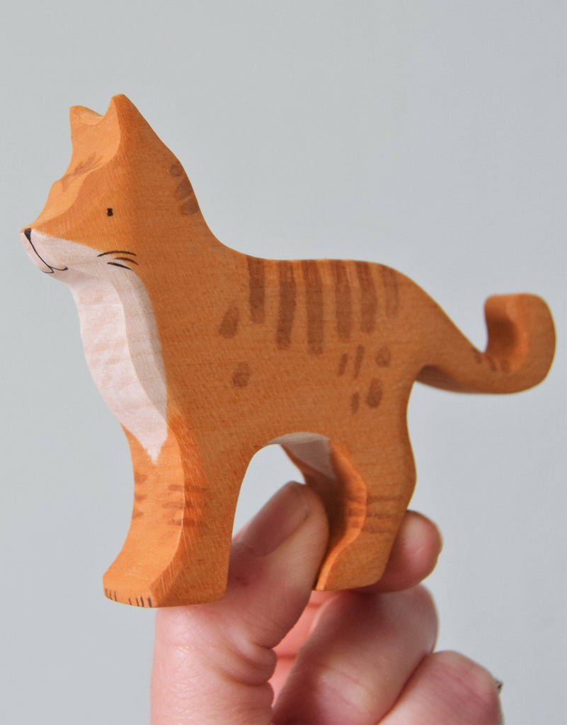 A hand holds the Handmade Wooden Ginger Cat, a small figurine with painted stripes, a white chest, and curled tail, crafted from sustainably sourced hardwood and finished with nontoxic plant-based stains.