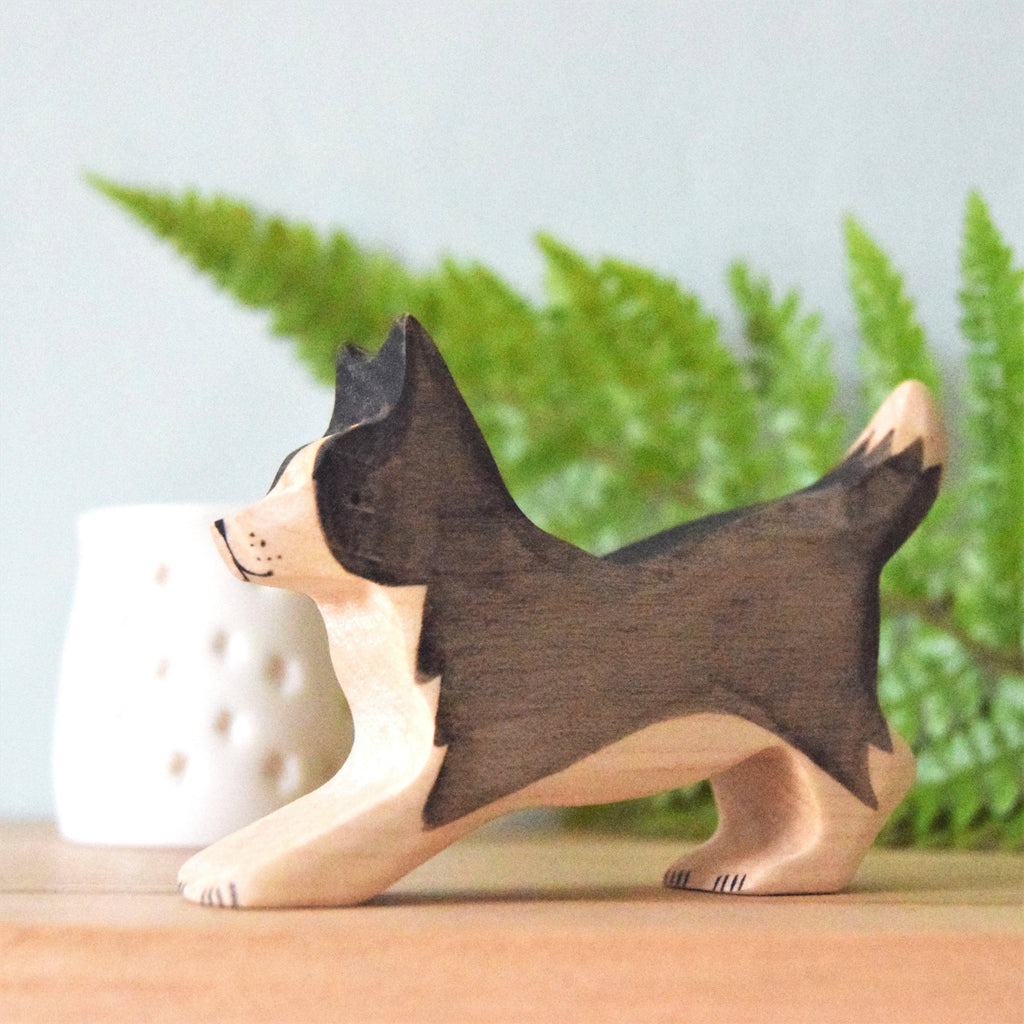 The Handmade Wooden Border Collie, crafted from sustainable hardwood and finished with natural plant-based stains, stands on a surface with a green fern and a small white decorative object blurred in the background.