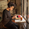 A person wearing headphones and sunglasses plays a Loog Piano with velocity-sensitive keys at a sunlit window-side counter in a cozy room, a coffee cup beside them.
