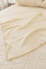 The Organic Pointelle Cotton Knit Baby Blanket, in a cream hue with a delicate lace edge, is neatly draped over a quilted bedspread in a softly lit room.