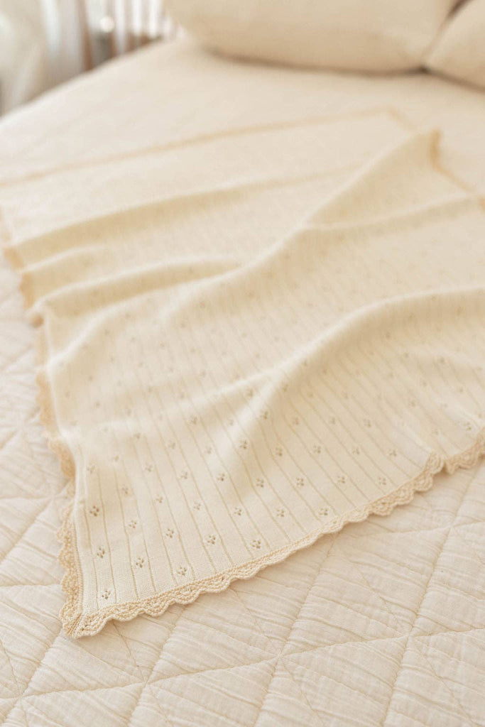 The Organic Pointelle Cotton Knit Baby Blanket, in a cream hue with a delicate lace edge, is neatly draped over a quilted bedspread in a softly lit room.