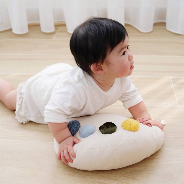 A baby in a white outfit lies on a light wooden floor, propped up on the Tummy Time Palette featuring colorful round patches, near a window with sheer white curtains.