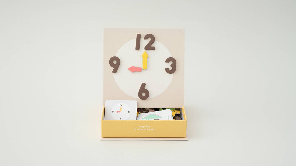 The Magnetic Number Play Set features a children’s magnetic clock toy with brown numbers and yellow and pink hands, shown above a yellow box filled with alphabet cards, magnet puzzles, and play pieces.