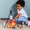 A young child sits on the floor playing with the Wooden Camp Fire Set, which includes a camp fire, pot, utensils, and food items under a tripod—ideal for indoor camping and imaginative pretend play.