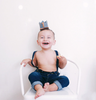 A smiling toddler wearing blue jeans, suspenders, and the Storm Gray Felt Crown sits barefoot on a white chair against a plain white background, ready for birthday celebrations.