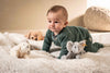 A baby in a green onesie crawls on a soft white blanket, surrounded by plush toys including the Steiff Floppy Linda Lamb 8-inch plush, an elephant, and a dog, with a textured cushion in the background.