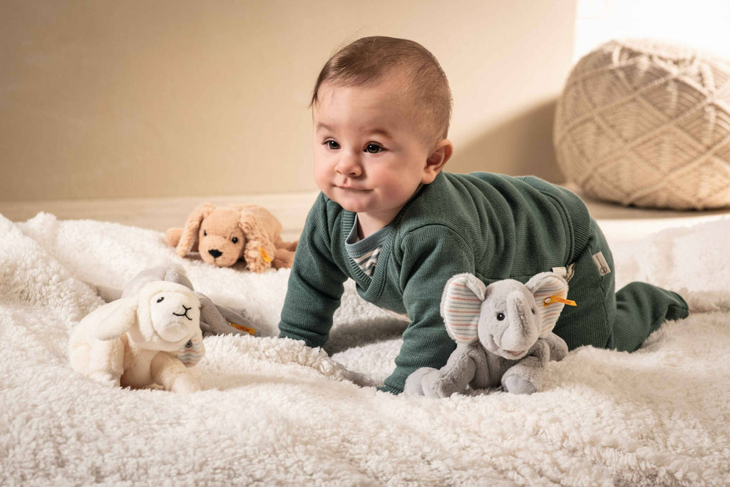 A baby in a green onesie crawls on a soft white blanket, surrounded by plush toys including the Steiff Floppy Linda Lamb 8-inch plush, an elephant, and a dog, with a textured cushion in the background.