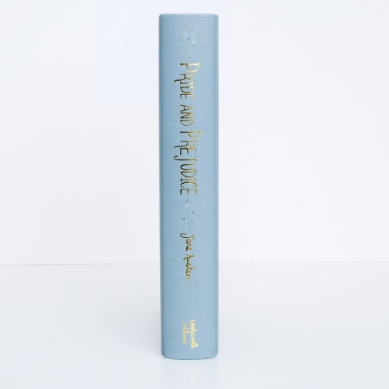 A light blue hardcover collector’s edition stands upright, its spine reading "Pride and Prejudice | Austen" in gold lettering with small decorative designs above and below the title.