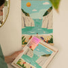 Someone holds the Diamond Painting Kit - Stiniva Beach by Maja Tomljanovic, showing a Mediterranean cove scene, while the finished artwork of cliffs, turquoise water, sailboats, and beachgoers hangs on the wall above.