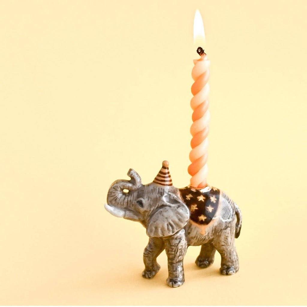 The Elephant Cake Topper is a fine porcelain figurine with a party hat and starry blanket, holding a lit orange candle—an adorable collectible keepsake perfect for celebrations, shown against a plain yellow background.