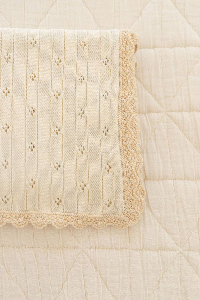 The Organic Pointelle Cotton Knit Baby Blanket, in folded cream, features a delicate eyelet pattern and scalloped edges, resting atop a quilted light beige surface.