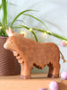 The Handmade Wooden Highland Cow, crafted from sustainable hardwood with carved details, is displayed on a wooden surface. In the background are a plant in a textured pot, string lights, and purple felt balls in the foreground.