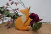 Handmade Wooden Snuggling Foxes, crafted from sustainable hardwood and finished with natural non-toxic stains, rest together on a tree stump accented by dried green leaves, pink flowers, and a purple rose on a plain light background.