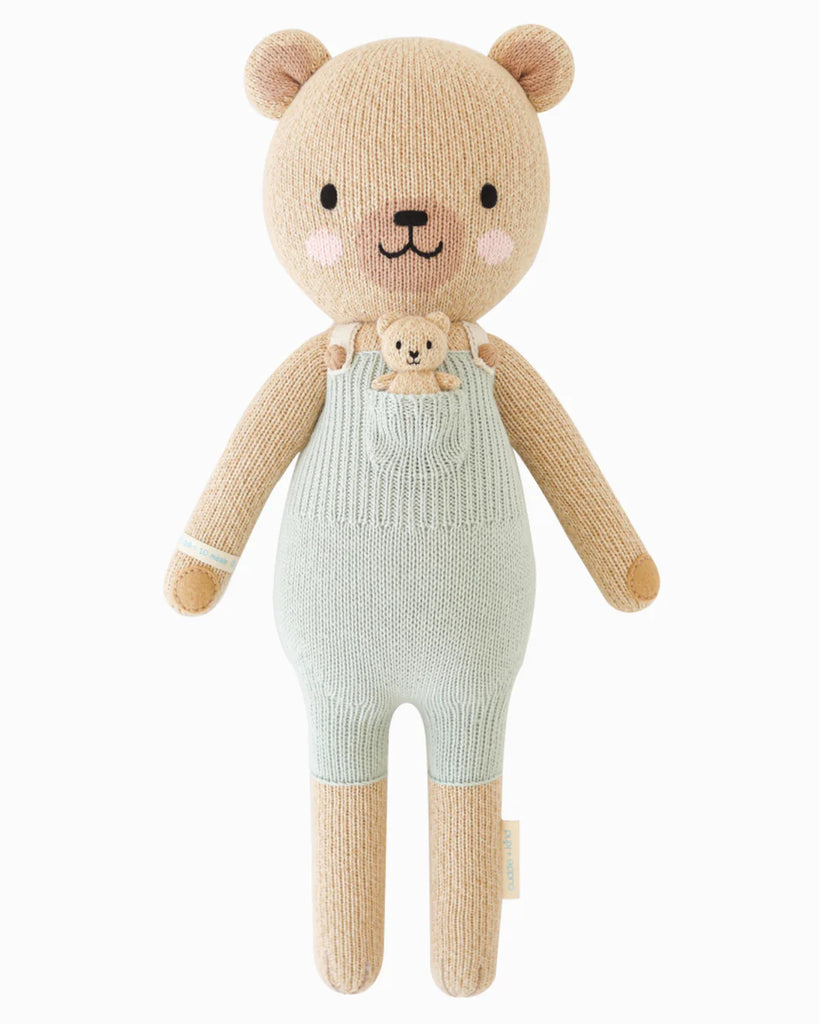 Cuddle + Kind Alexander The Honey Bear is a soft, knitted handmade teddy bear in light green overalls with a tiny bear in its pocket, filled with hypoallergenic polyfill and shown on a plain white background.