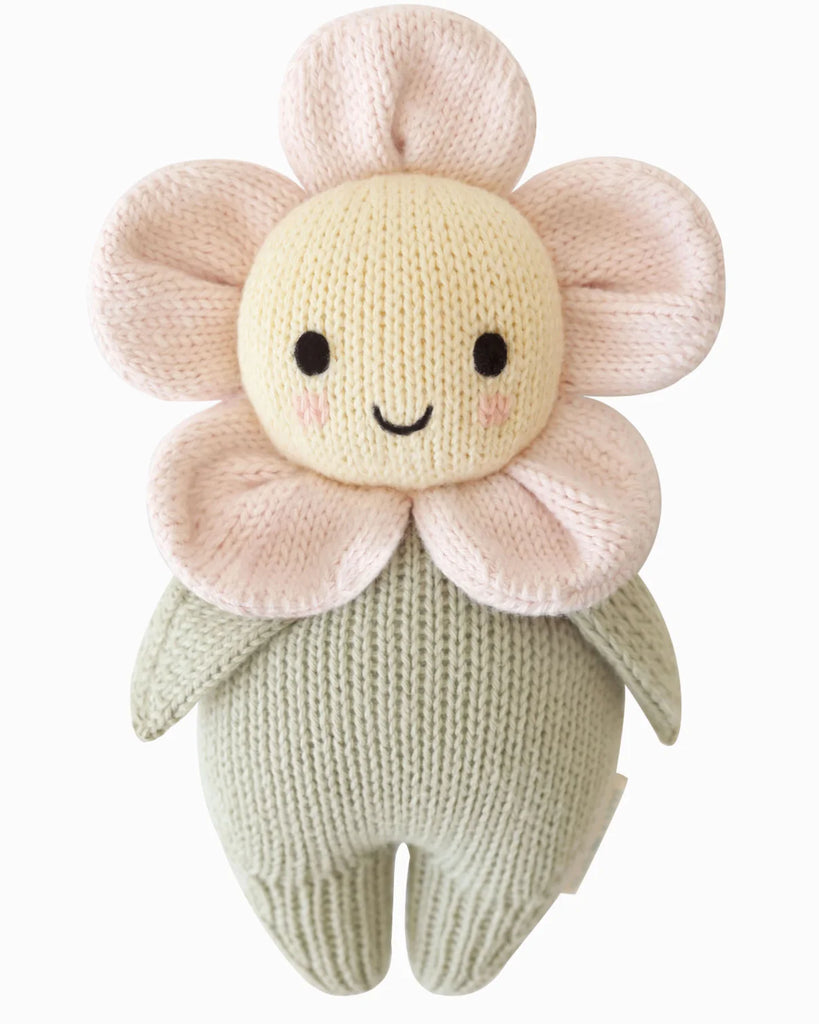 The Cuddle + Kind Baby Daisy - Blush Pink is a hand-knit plush flower toy with a smiling yellow face, pink petals, and a light green body, crafted from soft Peruvian cotton yarn.