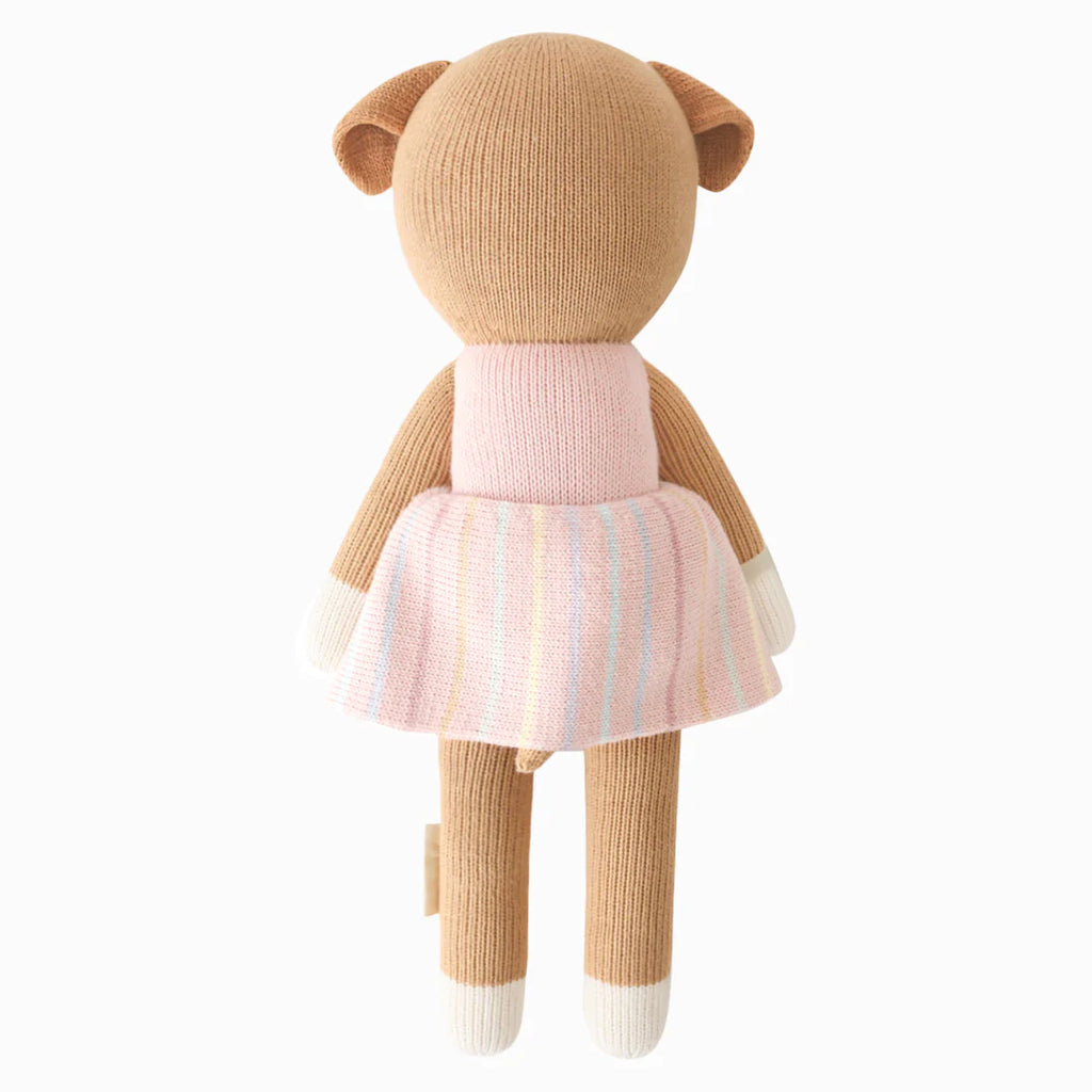 Cuddle + Kind Bea The Dog is a hand-knit stuffed animal, shown from the back. Made with brown yarn, white paws, and a light pink pastel-striped dress, and filled with hypoallergenic polyfill for extra comfort.