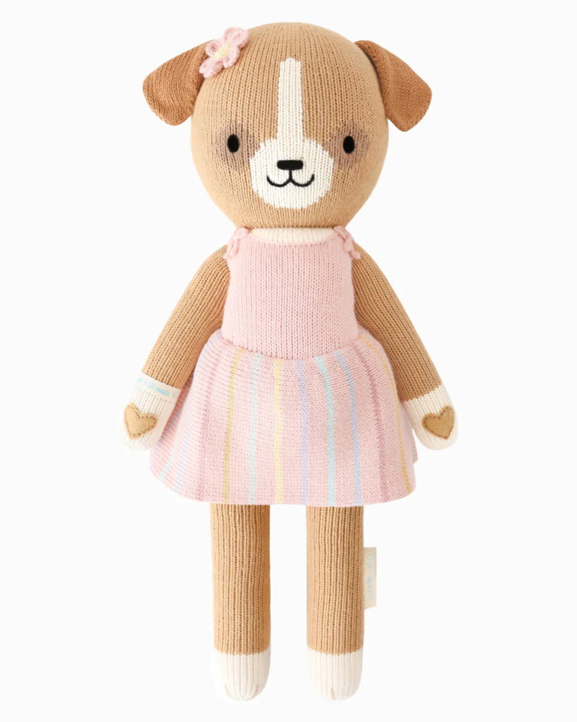 Cuddle + Kind Bea The Dog is a hand-knit stuffed doll with a beige body, white accents, and a smiling face. She wears a pink dress with pastel stripes and a pink flower on her head, and is filled with hypoallergenic polyfill.