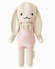 Cuddle + Kind Briar The Bunny - Peony Pink is a hand-knit, fair trade bunny doll with long floppy ears, pink overalls, a small matching bunny in the pocket, embroidered details, and a soft plush design.