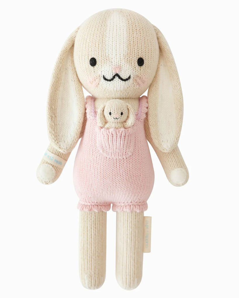 Cuddle + Kind Briar The Bunny - Peony Pink is a hand-knit, fair trade bunny doll with long floppy ears, pink overalls, a small matching bunny in the pocket, embroidered details, and a soft plush design.