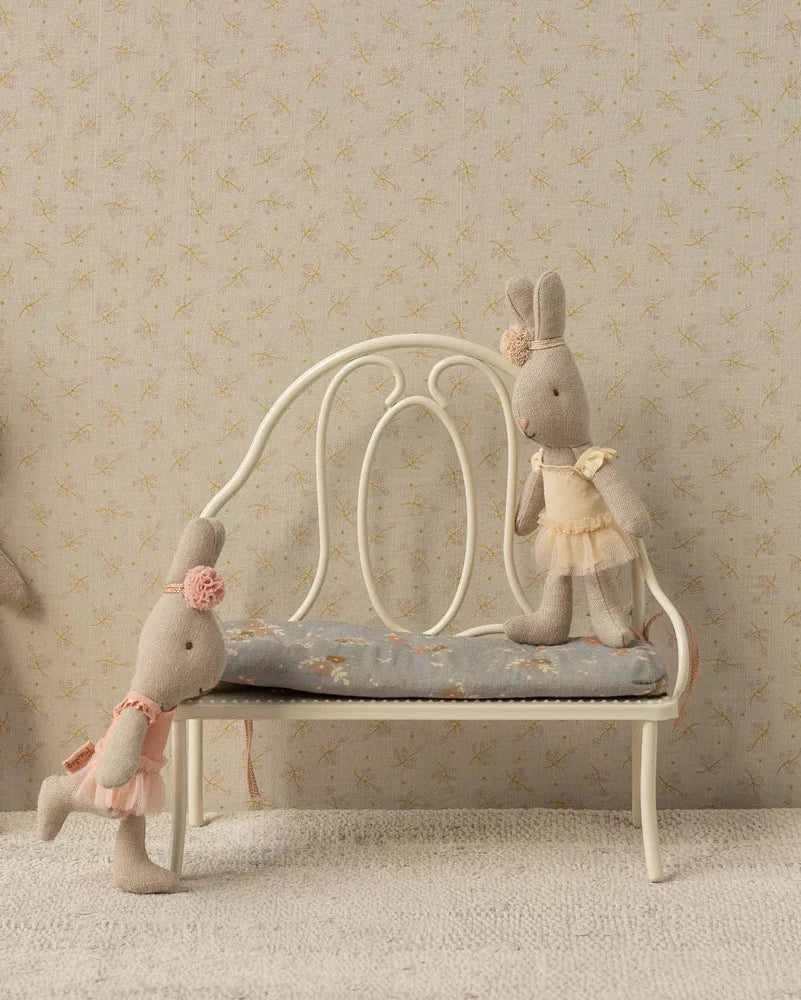 Three fabric bunny dolls in pastel dresses pose with the Maileg Vintage Bench, Miniature - Off White: one stands, one sits comfortably on the bench, and another leans playfully with its arms on it.