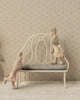Three fabric bunny dolls in pastel dresses pose with the Maileg Vintage Bench, Miniature - Off White: one stands, one sits comfortably on the bench, and another leans playfully with its arms on it.