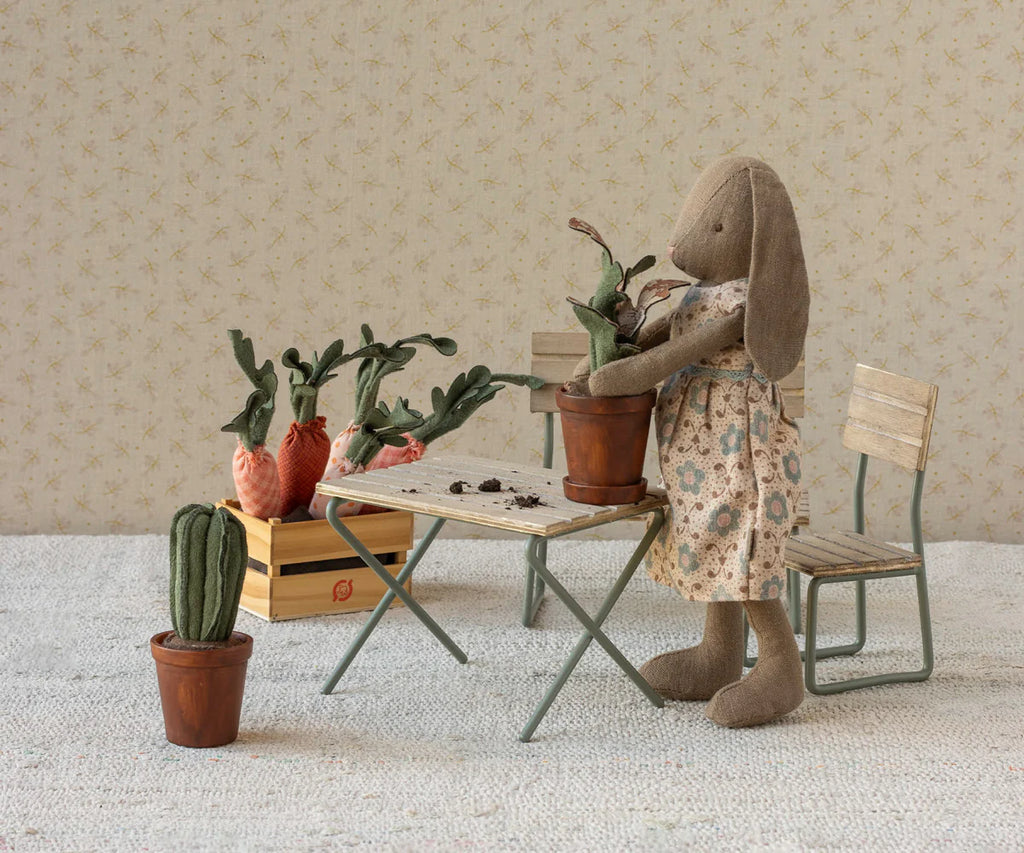 A stuffed bunny in a floral dress stands by a table potting a fabric plant. More greenery fills a crate on a bench, and the Maileg Flower Pot With Cactus, Miniature sits on the floor amid vintage-style décor.