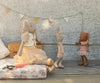 A Maileg Bunny, Size 1, in chocolate brown wears a rose ballet suit and skirt, posed like a dancer on a floral mattress with suitcases behind and pastel bunting on the textured wall.