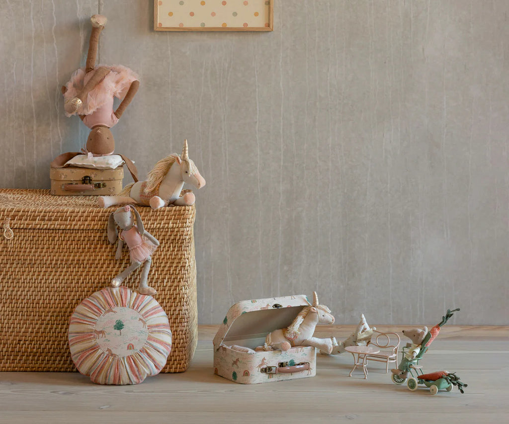 A woven basket with stuffed animals, a striped round pillow, a doll, unicorn-themed toys, and the Maileg Suitcase Set, 2-Pieces - Unicorn for toy storage are displayed on a light wooden floor against a gray wall with a framed picture above.