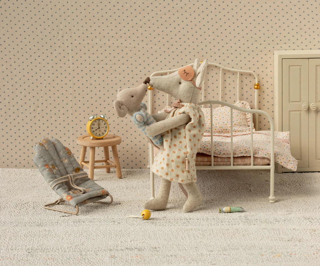 A Maileg Mom Mouse in a nightgown (Maileg Extra Clothing: Nightgown For Mom Mouse) cuddles her baby mouse in a pastel-toned miniature bedroom with bed, chair, yellow clock, and small toys scattered on the floor.