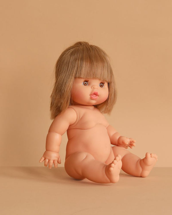 The Minikane Baby Doll (13") - Zoe sits on a beige surface against a matching background, featuring straight light brown hair, wide eyes, parted lips, chubby limbs, and lifelike details. Clothing sold separately.