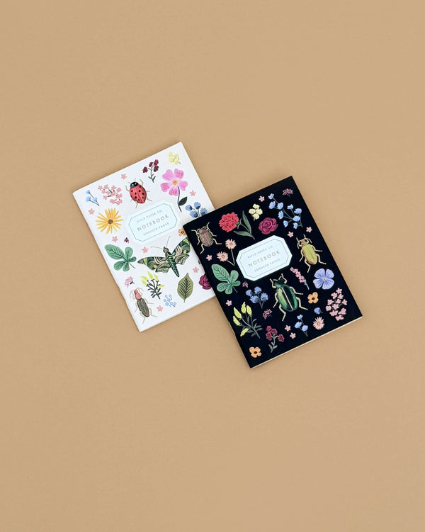 A Pair of 2 Curio Pocket Notebooks, featuring botanical and insect cover illustrations—one white and one black—lie slightly overlapping on a beige background. Each notebook measures 10.8 x 14 x 1 cm (4.3 x 5.5 x 0.4 in).