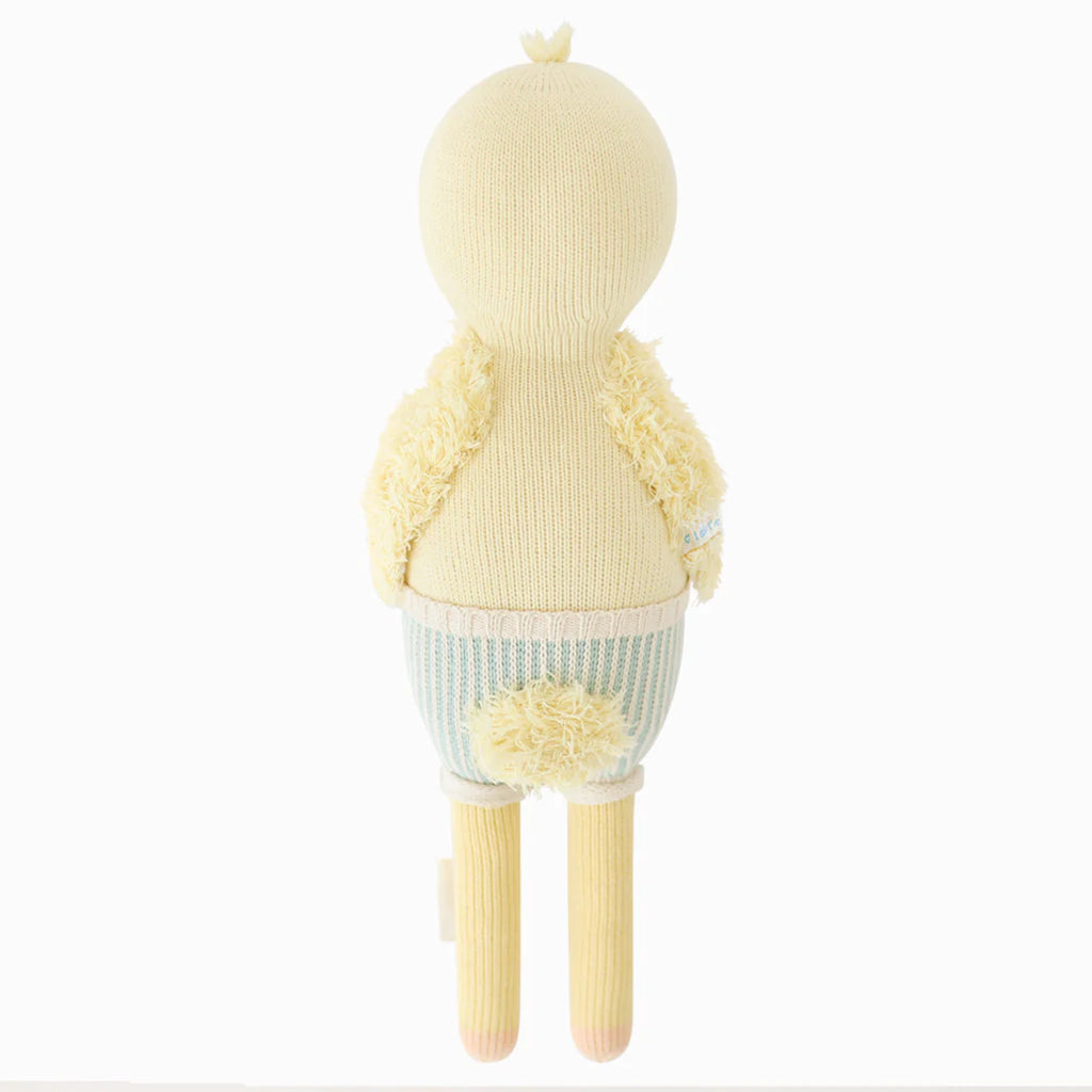The Cuddle + Kind Duckling is a hand-knit yellow plush toy with a round head, fuzzy wings, small tail, long striped legs, and blue and white shorts, shown from the back against a plain white background.