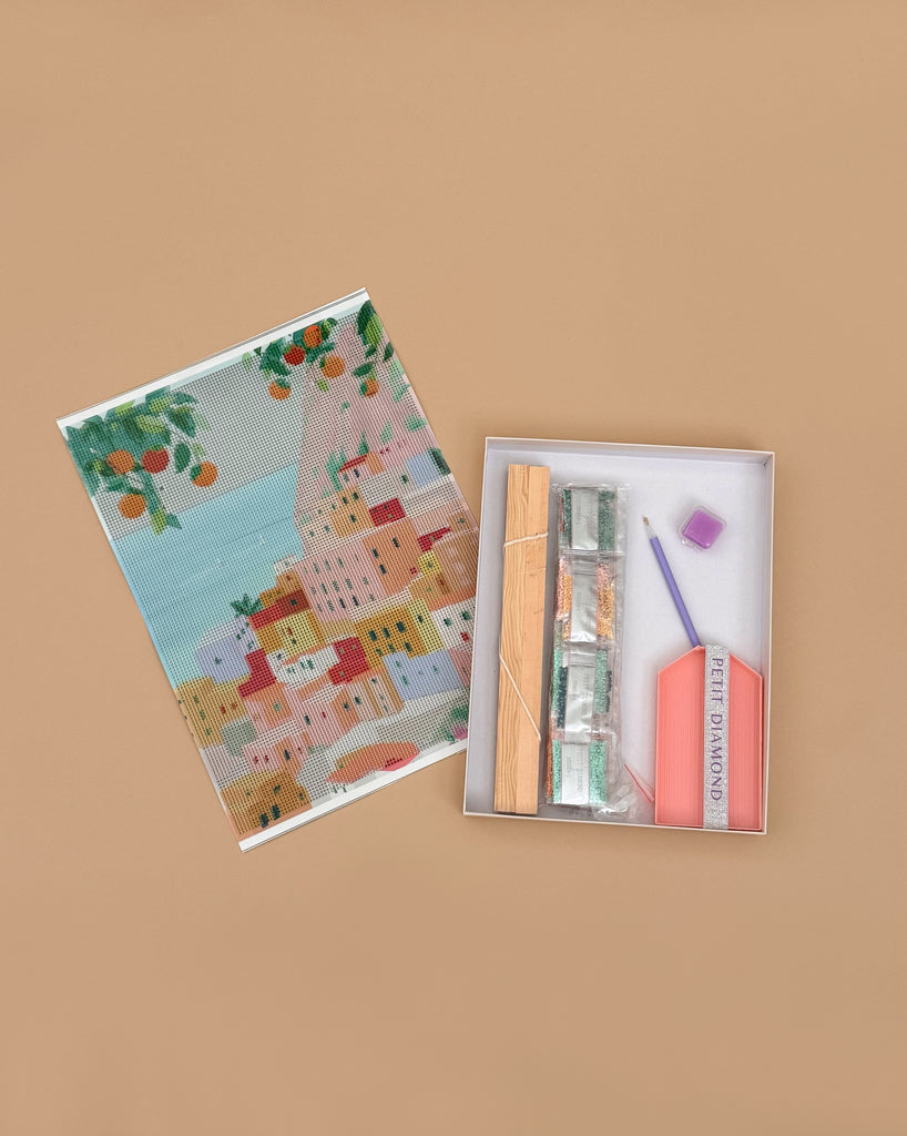 A flat lay of the Diamond Painting Kit - Positano by Petra Lidze on a tan background, featuring a colorful canvas of houses and trees, bags of gems, and all tools neatly arranged in an open box.