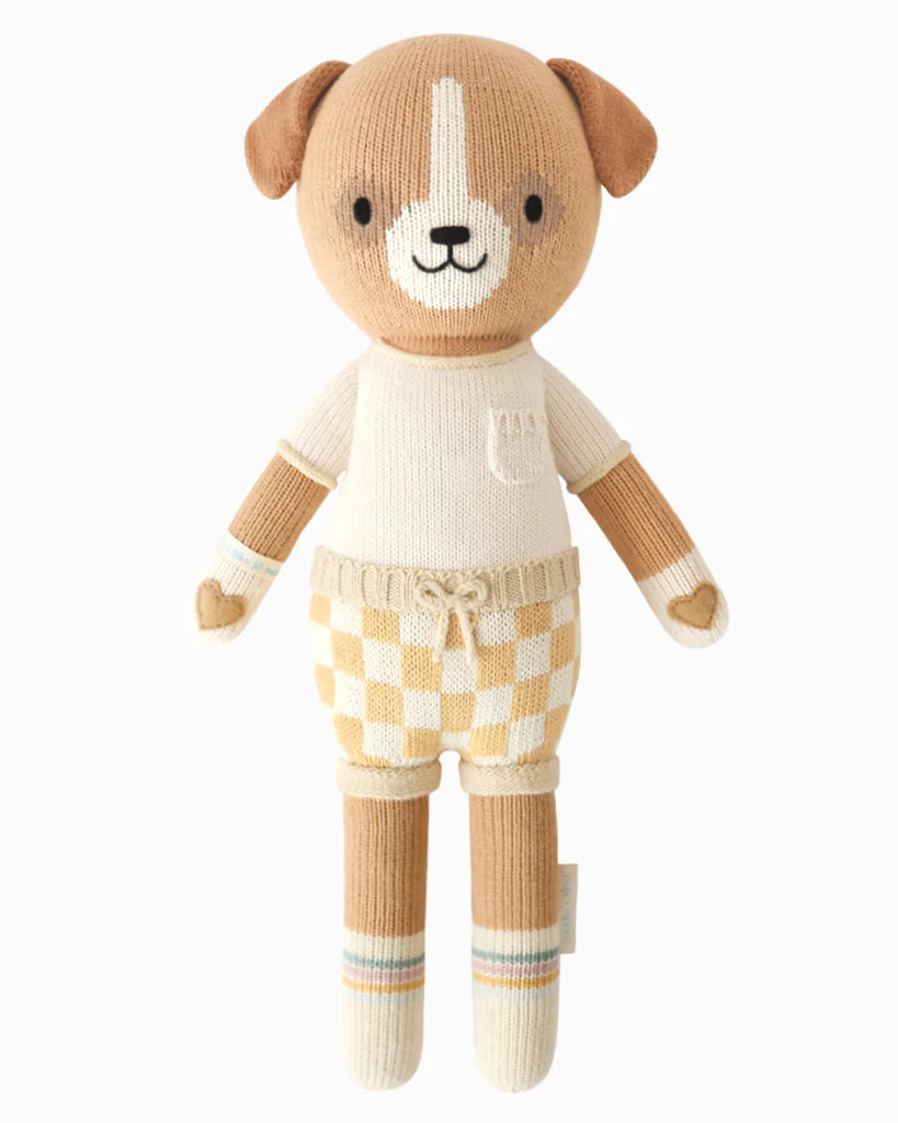 Cuddle + Kind George The Dog is a hand-knit brown and cream doll dog toy, wearing a cream shirt with pocket and beige checkered shorts. This smiling pup has floppy ears, heart-shaped hands, and is filled with hypoallergenic polyfill.