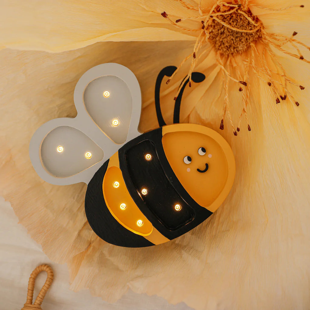 The Handmade Wooden Mini Lamp - Happy Bee is a cute, bee-shaped night light with glowing LED wings and body, resting on a pale yellow flower petal. Its smiling black-and-yellow design adds charm to any kids' room decor.